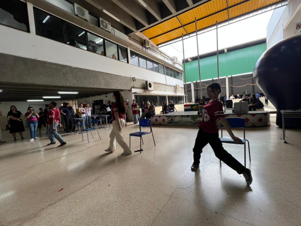 Uma foto de uma área interna ampla e coberta, mostrando jovens em movimento. Duas jovens no primeiro plano, vestindo camisas esportivas vermelhas e brancas, estão em poses de dança ou coreografia, com cadeiras de escola ao redor. Outras pessoas assistem ao fundo. O piso é claro e liso.