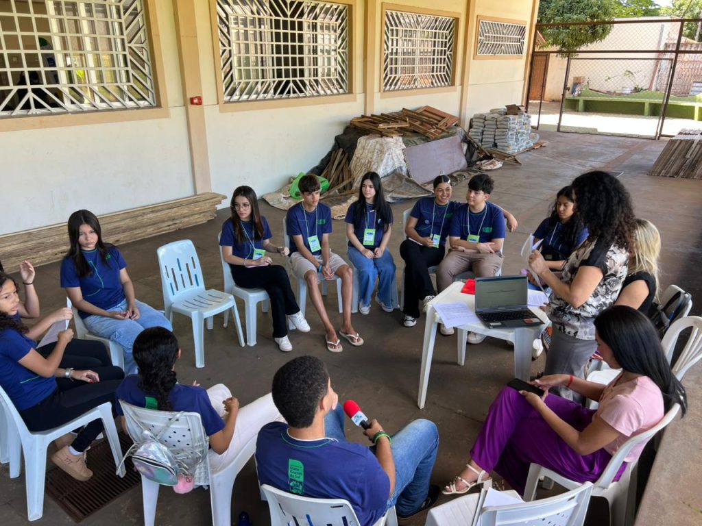 Um grupo de jovens e duas mulheres adultas estão reunidos em um círculo, sentados em cadeiras plásticas brancas, em uma área externa coberta. Uma mulher está de pé no centro, lendo ou instruindo o grupo. Há uma mesa baixa com um laptop, pastas e papéis no centro da roda.