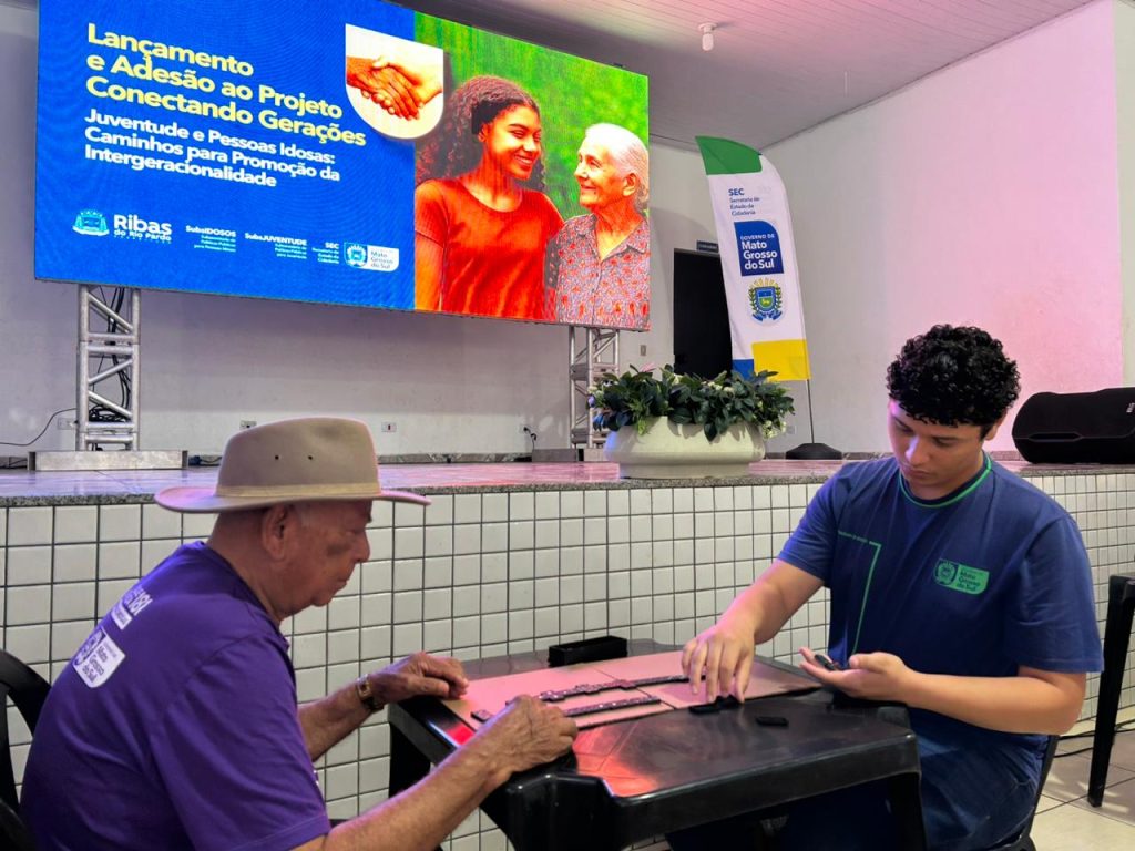 Em primeiro plano, um homem idoso, vestindo um chapéu e uma camiseta roxa, e um jovem, em uma camiseta azul, estão sentados em uma mesa escura jogando dominó. Ao fundo, uma grande tela de projeção (telão) exibe o tema do evento: "Lançamento e Adesão ao Projeto Conectando Gerações: Juventude e Pessoas Idosas: Caminhos para Promoção da Intergeracionalidade".