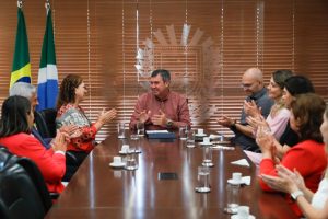 Vista frontal da mesa. O homem de camisa avermelhada está sentado à cabeceira, sorrindo e batendo palmas. As outras pessoas ao redor da mesa também aplaudem em um gesto de celebração ou concordância. Ao fundo, uma persiana de madeira com o brasão do Estado de Mato Grosso do Sul gravado no vidro e as bandeiras do Brasil e do Estado à esquerda.