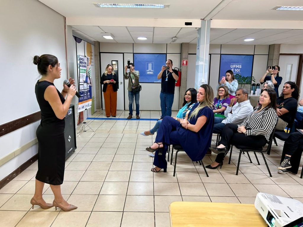 Uma mulher vestida de preto, em pé à esquerda, está falando em um microfone de mão para uma plateia sentada em cadeiras escuras. O público, que inclui fotógrafos profissionais e outros participantes, observa a apresentação. A sala é clara, com portas e cartazes de fundo.