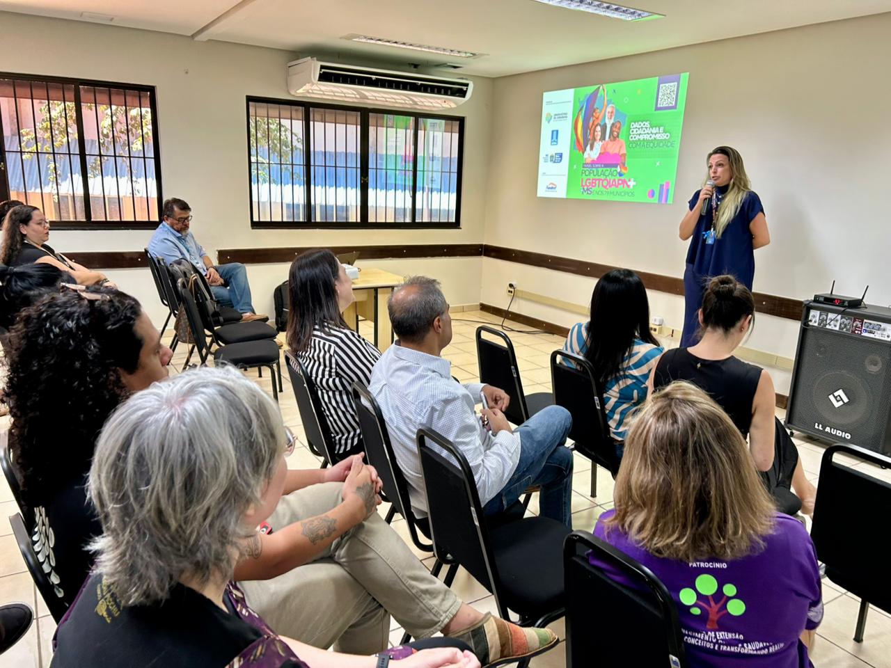 ista do público assistindo a uma apresentação. A oradora (uma mulher de blusa azul marinho) está à direita, em pé, falando em frente a um telão. O telão mostra gráficos e textos relacionados a "DADOS CIDADANIA E DIREITOS HUMANOS" e "PAINEL LGBTQIA+". O público, com várias idades e estilos, ouve atentamente.