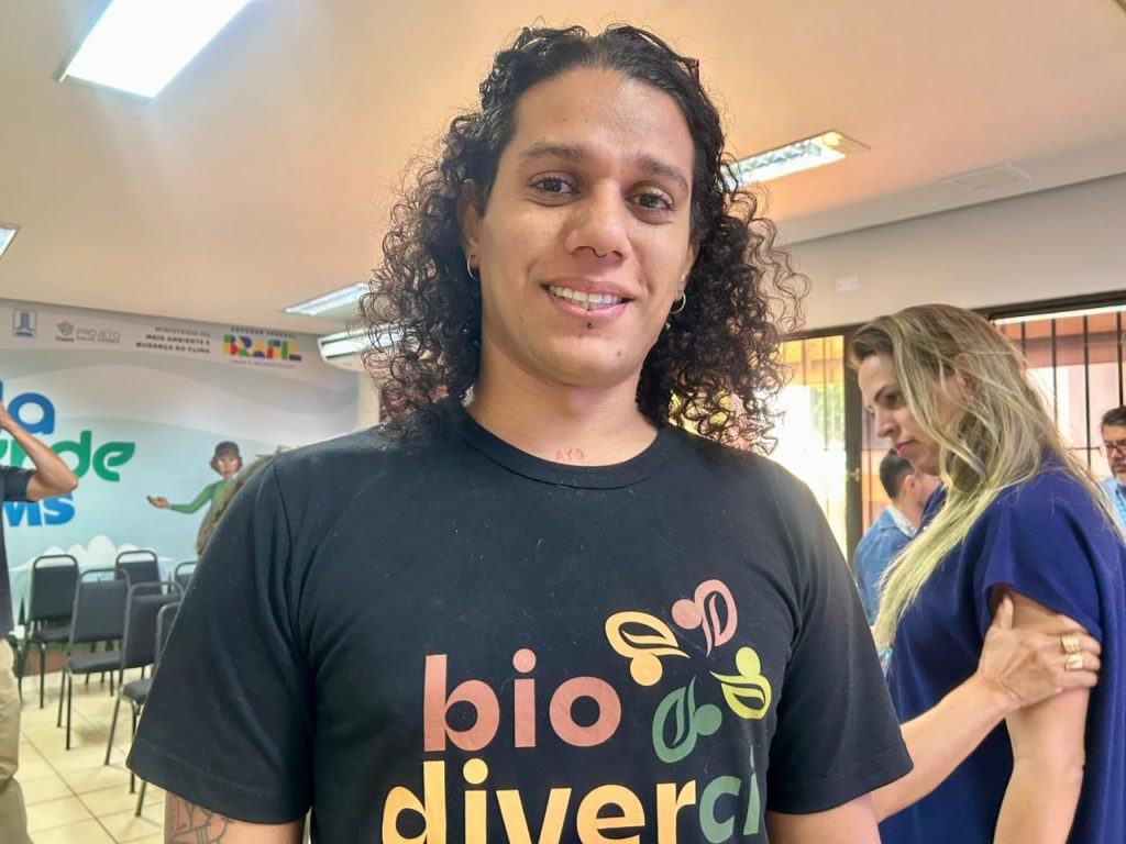 Close de uma jovem sorrindo, com cabelo cacheado e longo. Ela veste uma camiseta preta com a palavra "bio diversa" escrita em cores vibrantes (laranja, verde, amarelo). Em segundo plano, uma mulher de blusa azul marinho e um homem de camisa azul estão visíveis.