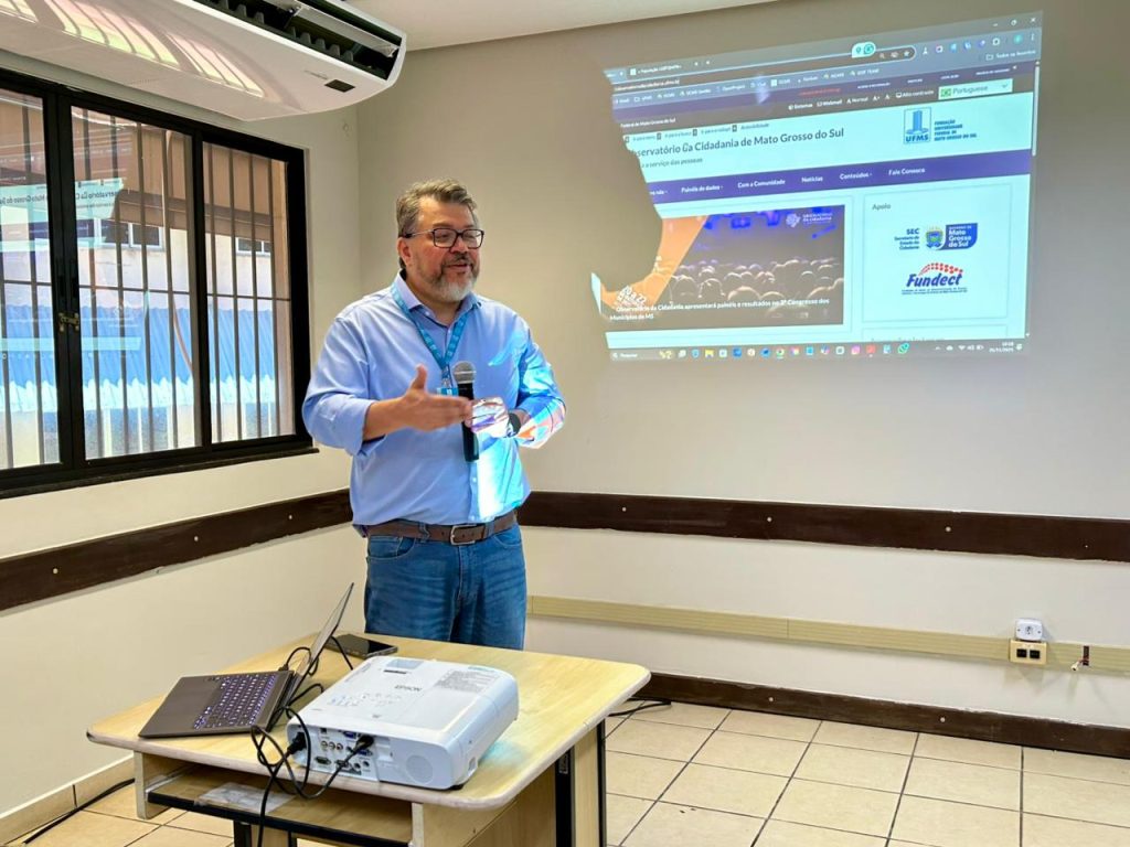 Um homem barbudo de camisa social azul clara e jeans está em pé, no centro, apresentando. Ele gesticula com a mão direita em direção a um telão projetado na parede, que exibe a página principal do "Observatório da Cidadania de Mato Grosso do Sul" e logotipos de apoio (SEC, Governo MS, Fundect). Um projetor e um notebook estão sobre uma mesa no primeiro plano.