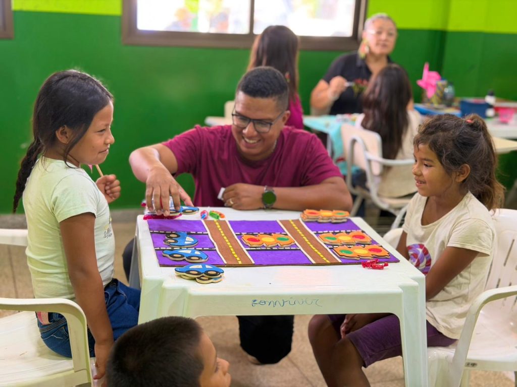 Um homem de camiseta bordô sorri ao interagir com duas crianças sentadas à sua frente em uma mesa branca, participando de um jogo educativo de trânsito com carrinhos e semáforos de papel.
