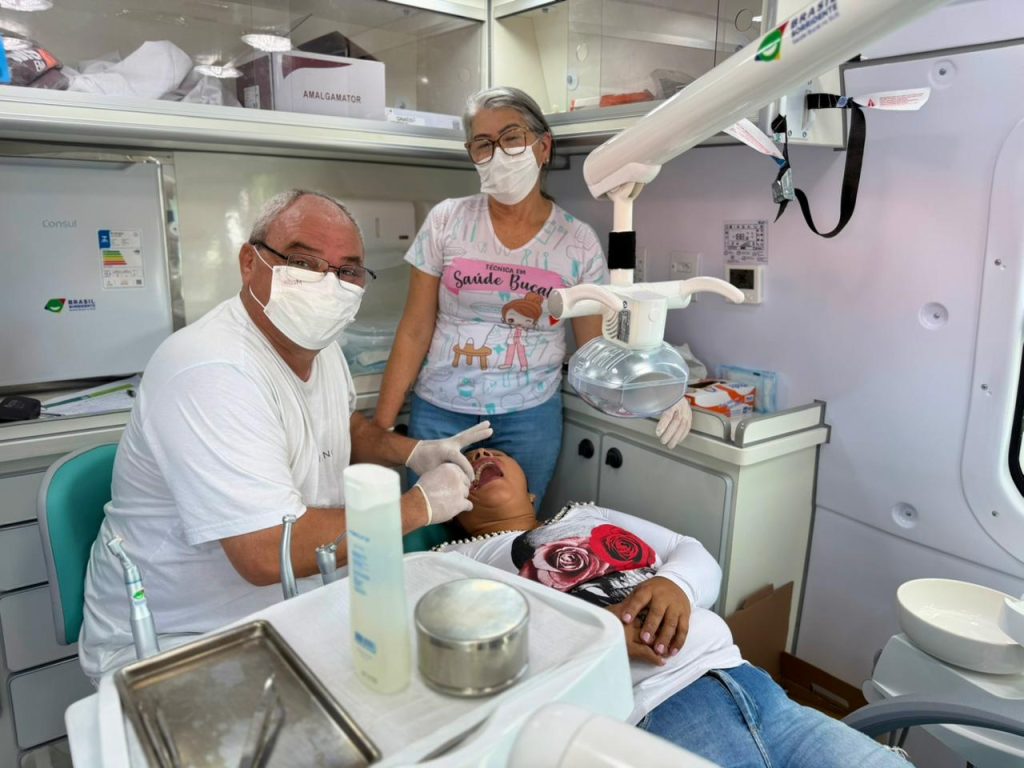 Cena de atendimento de saúde em uma unidade móvel (ônibus ou van). Um dentista de máscara e jaleco branco examina a boca de uma mulher deitada na cadeira. Uma assistente de máscara e camisa estampada está ao lado.