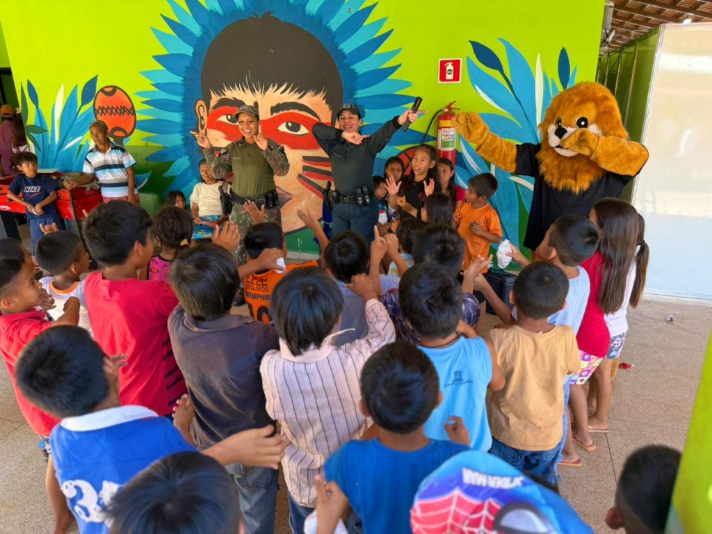 Uma cena vibrante ao ar livre em frente a uma parede pintada com um mural indígena colorido. Duas policiais sorridentes interagem com um grande grupo de crianças que as cercam. Um mascote com fantasia de leão marrom está à direita.