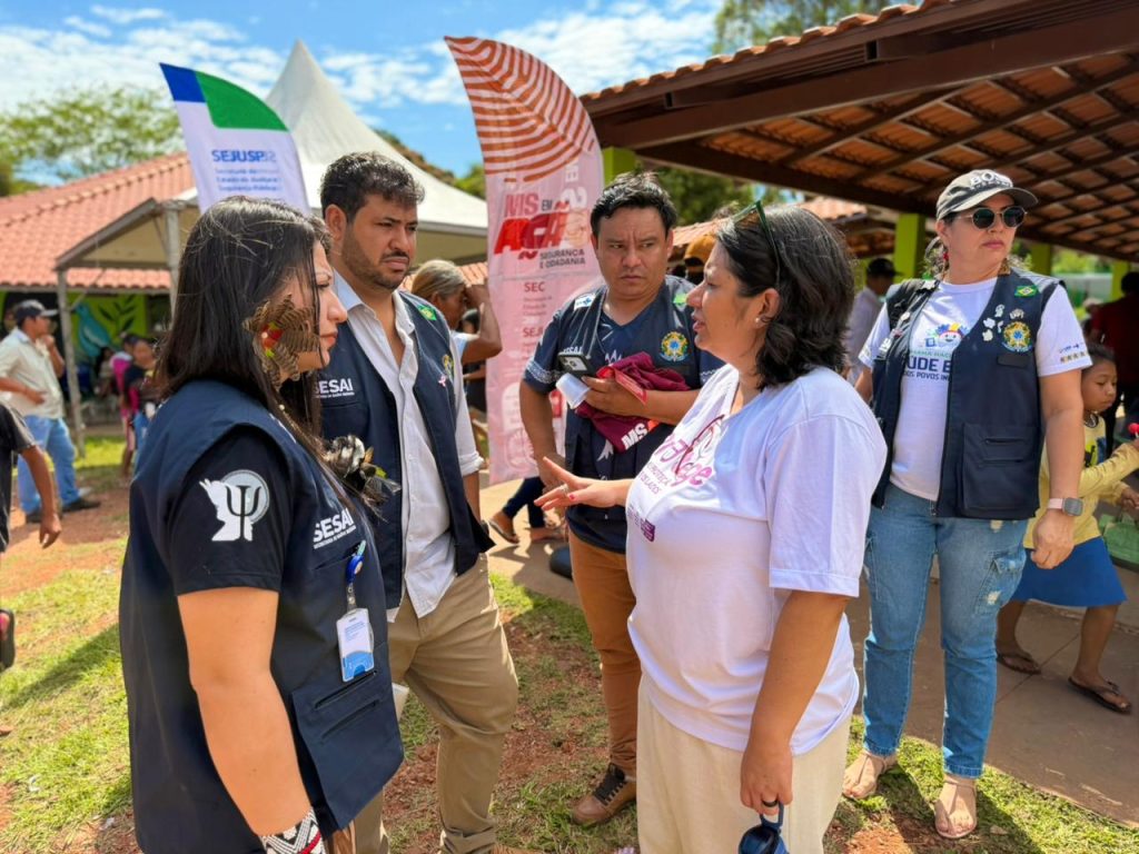 Quatro pessoas, incluindo duas mulheres e dois homens, estão em uma conversa informal ao ar livre. Três deles vestem coletes escuros (Sesai/SESAU) com logotipos de serviços. Uma das mulheres (à direita) de camiseta branca e a outra (à esquerda) com adereços indígenas interagem com os homens. Bandeiras de divulgação dos programas MS em Ação e SEJUSP estão visíveis ao fundo.