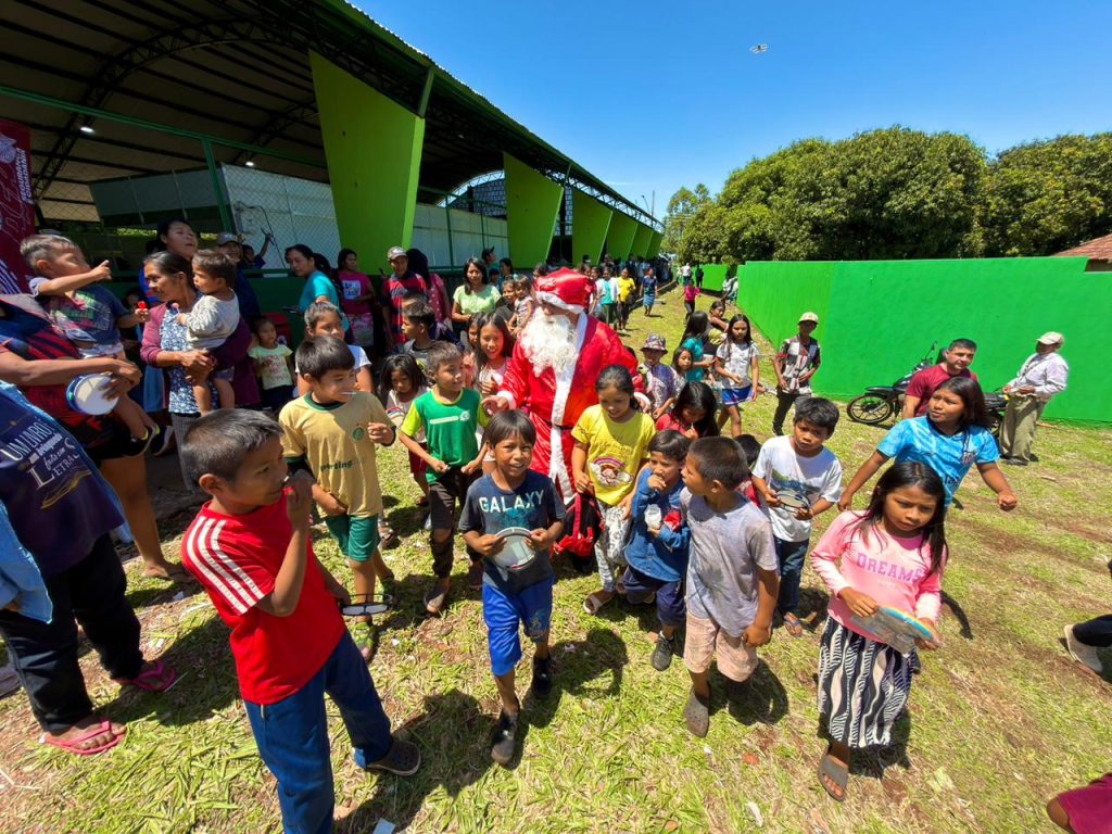 Uma foto que mostra a chegada do Papai Noel em uma área gramada e aberta. Ele está cercado por dezenas de crianças, que o olham curiosas e animadas. Ao fundo, há uma estrutura coberta com paredes verdes.