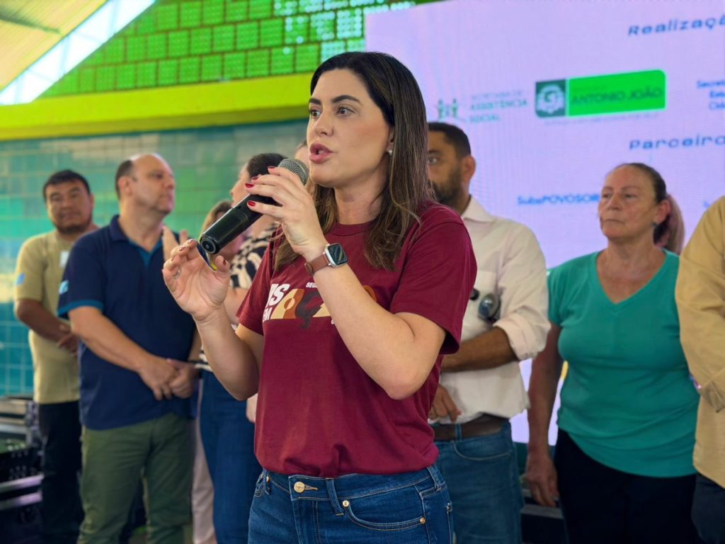 Uma mulher de camiseta bordô escura e calça jeans fala ao microfone, em um palco. Ela está cercada por outras pessoas (servidores e autoridades) que a observam.