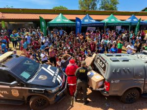 Uma visão aérea que mostra Papai Noel e dois policiais militares (DOF - Departamento de Operações de Fronteira) ajudando a descarregar caixas de presentes da caçamba de uma caminhonete policial. Uma caminhonete da Polícia Civil também está estacionada. Uma grande multidão de crianças e adultos está reunida ao fundo, sob tendas da UEMS (Universidade Estadual de Mato Grosso do Sul).