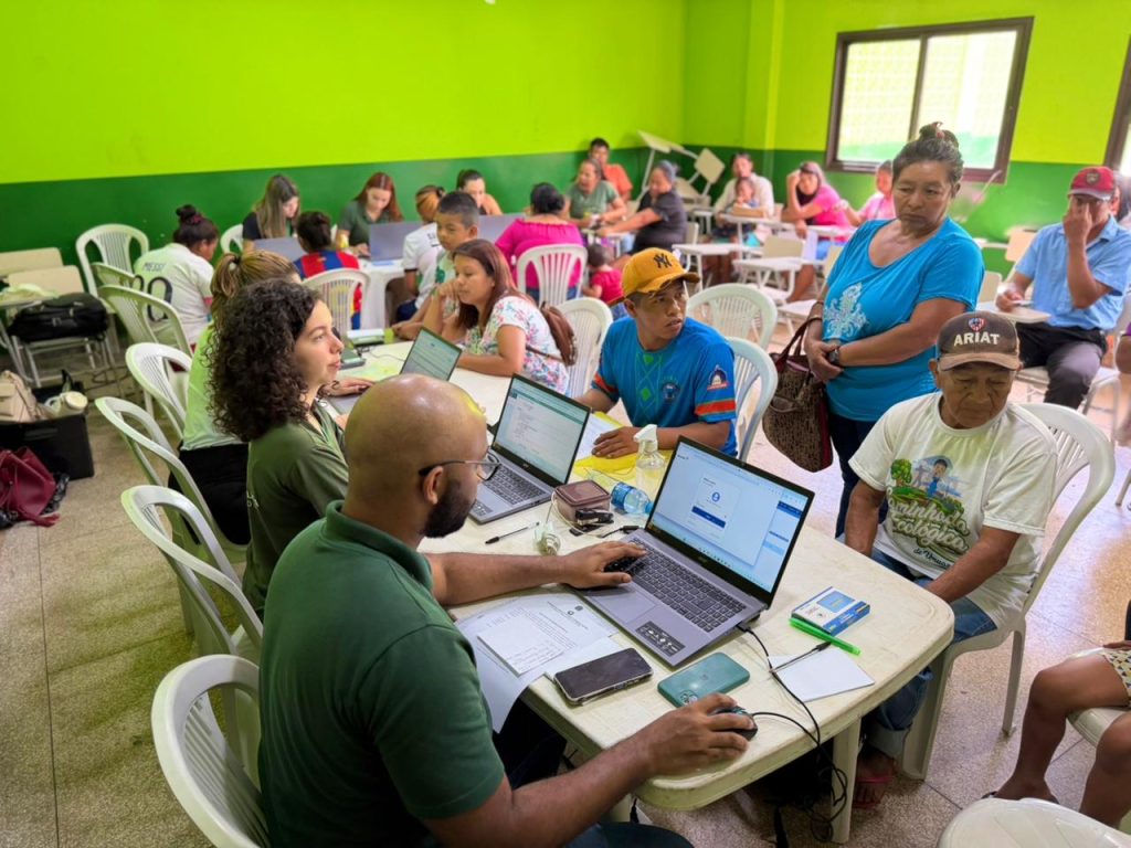 Um ambiente interno de sala de aula com paredes verdes e mesas brancas. Servidores e voluntários, usando laptops, ajudam membros da comunidade (incluindo adultos e idosos) com cadastros ou acesso a serviços digitais.