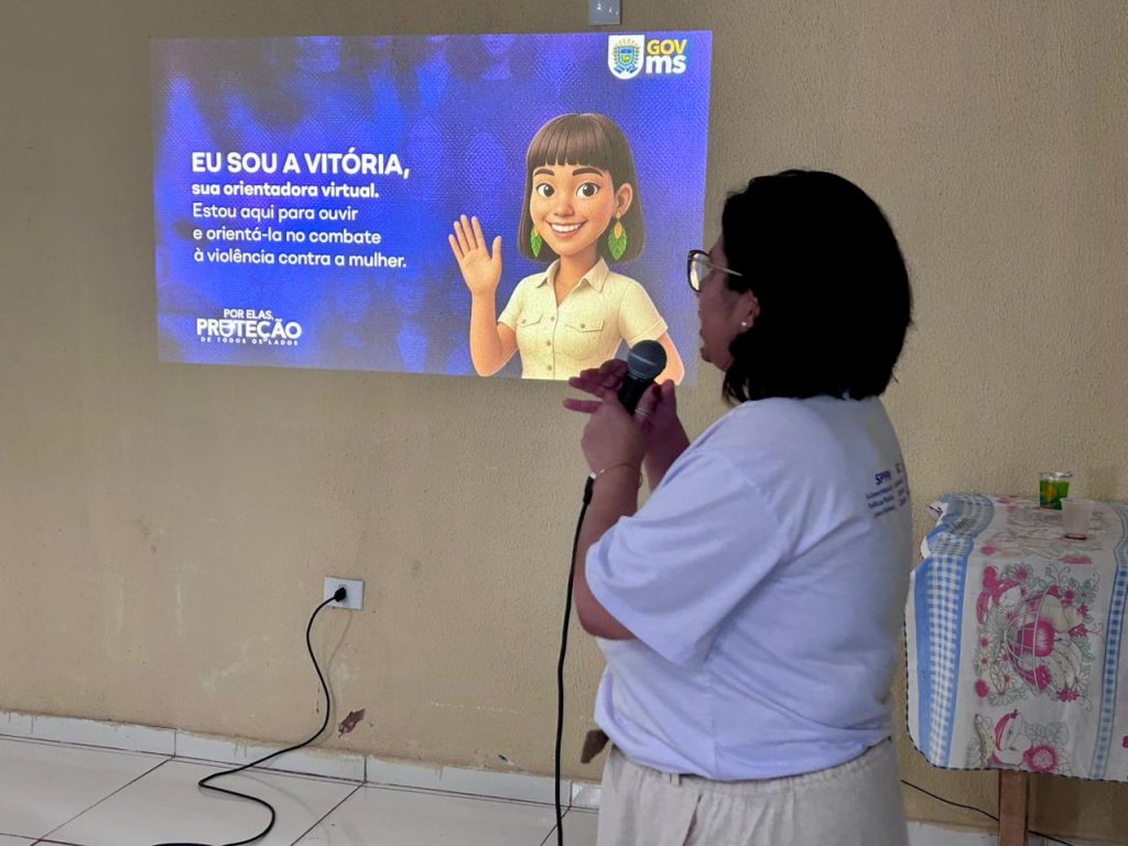 Uma mulher, vista de costas, está falando em um microfone de mão em frente a uma parede clara. Uma imagem é projetada na parede, mostrando uma personagem de desenho animado chamada "EU SOU A VITÓRIA, sua orientadora virtual", parte de uma campanha de combate à violência contra a mulher do Gov MS.