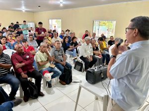 A foto mostra um homem, visto de costas em primeiro plano, falando em um microfone para uma audiência grande e densamente reunida de pessoas mais velhas e jovens, em um salão simples (Evento PERIFEIRARTE CAMPO GRANDE).