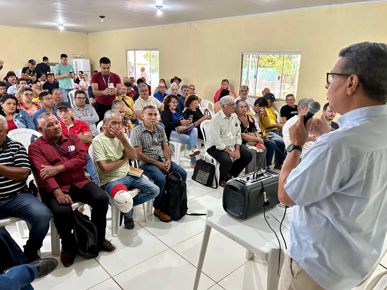 A foto mostra um homem, visto de costas em primeiro plano, falando em um microfone para uma audiência grande e densamente reunida de pessoas mais velhas e jovens, em um salão simples (Evento PERIFEIRARTE CAMPO GRANDE).