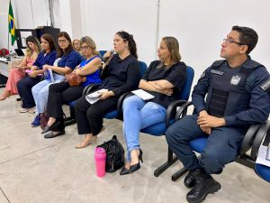 Close de uma fileira de participantes sentados em cadeiras azuis. A maioria é mulher, com uma delas (de blusa preta) falando em um microfone de mão. Na extrema direita, um homem vestido com um colete balístico sobre o uniforme da Polícia Militar (PMMS) está sentado com as mãos sobre as pernas, observando o orador.