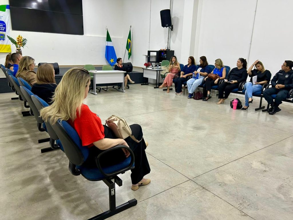 Vista ampla de uma sala de reuniões. Os participantes estão sentados em cadeiras azuis dispostas em um semicírculo aberto, com a maioria das pessoas concentradas no fundo e nos lados. Uma mulher de blusa vermelha é vista de costas em primeiro plano. Ao fundo, perto das bandeiras do Brasil e de Mato Grosso do Sul, há uma mesa com equipamentos eletrônicos.