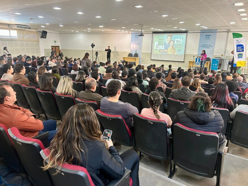 Fotografia tirada da parte de trás de um auditório lotado. Dezenas de pessoas estão sentadas em poltronas pretas com detalhes vermelhos, assistindo a uma apresentação. No palco ao fundo, há um telão projetando o slide do programa “Juventude Plena”. À direita do palco, uma mulher fala em um púlpito com o logo da UFMS e do Governo do Estado de Mato Grosso do Sul. Um intérprete de Libras está posicionado à esquerda do telão.