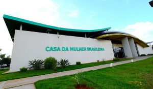Foto de ângulo aberto da fachada externa de um prédio moderno branco. A estrutura tem um design curvo e um telhado verde. O nome "CASA DA MULHER BRASILEIRA" está escrito em letras grandes e verdes na parede. Há gramado e vegetação na frente.