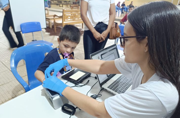 Mulher com luvas azuis auxiliando uma criança, sentada em uma cadeira azul, a usar um aparelho de scanner em uma mesa branca com um notebook. A cena mostra atendimento de serviço público.