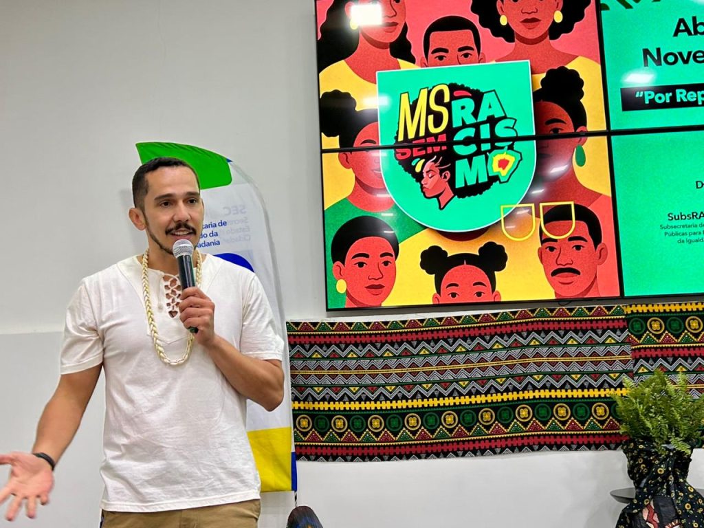 Fotografia de um homem jovem, de pele parda e barba curta, falando ao microfone em um evento. Ele veste uma camiseta branca e um colar de contas claras. Ao fundo, um grande telão exibe a identidade visual do evento com o título "Abertura do Novembro Negro - Por Reparação e Bem Viver" e a logo "MS SEM RACISMO", ilustrada com rostos de pessoas negras em estilo gráfico colorido.