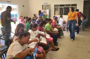 Foto do interior de uma sala de espera durante um mutirão de atendimento. À esquerda e ao centro, várias pessoas, incluindo homens e mulheres indígenas, estão sentadas em filas segurando pastas e documentos. À direita, uma mulher com camiseta amarela caminha pelo corredor. Ao fundo, vê-se uma porta com a placa "Secretaria" e uma funcionária organizando um banner. O ambiente é bem iluminado e possui paredes claras.