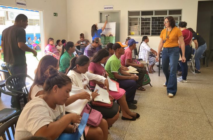 Foto do interior de uma sala de espera durante um mutirão de atendimento. À esquerda e ao centro, várias pessoas, incluindo homens e mulheres indígenas, estão sentadas em filas segurando pastas e documentos. À direita, uma mulher com camiseta amarela caminha pelo corredor. Ao fundo, vê-se uma porta com a placa "Secretaria" e uma funcionária organizando um banner. O ambiente é bem iluminado e possui paredes claras.