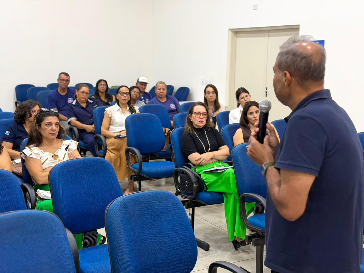 Fotografia tirada de trás do palestrante, mostrando sua silhueta de costas em primeiro plano à direita, segurando o microfone. A imagem foca na plateia à sua frente, composta por cerca de 15 a 20 pessoas sentadas em poltronas azuis dispostas em fileiras. Os participantes, homens e mulheres de diversas idades, demonstram atenção e alguns fazem anotações. O ambiente é amplo, com paredes brancas e piso de cimento queimado claro.