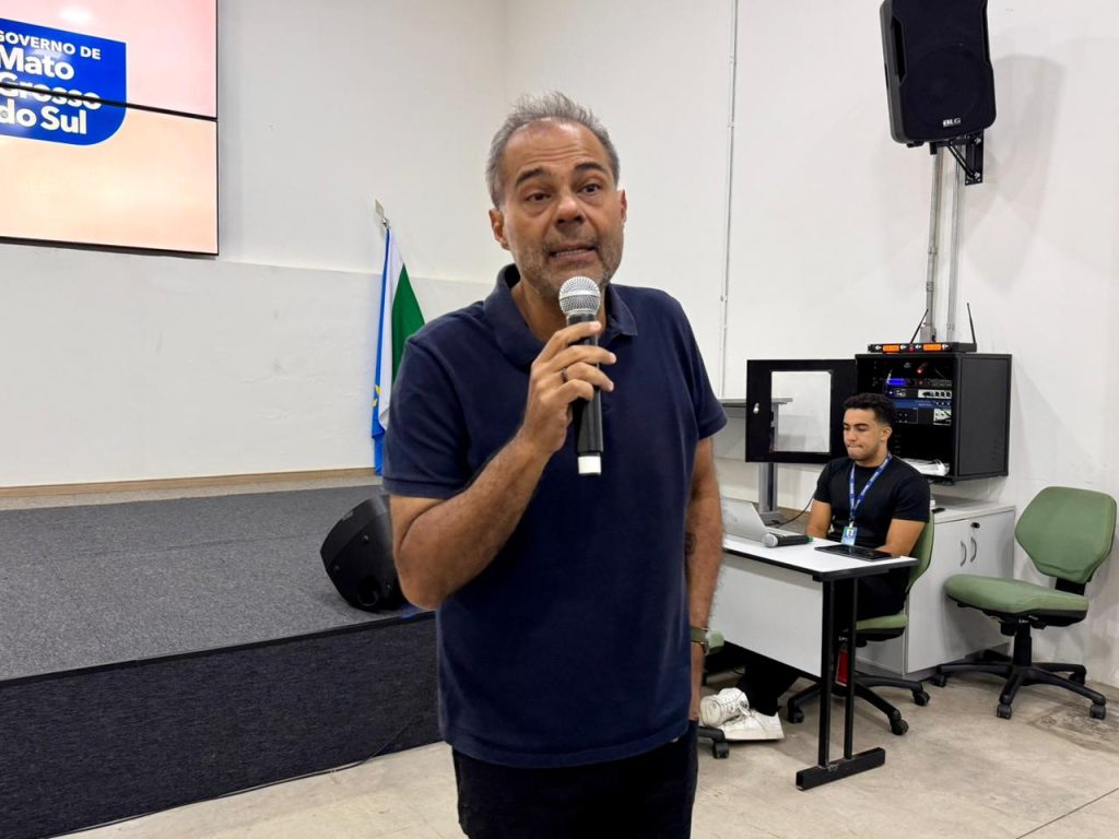 Fotografia em plano médio de um homem de meia-idade, de pele parda e cabelos grisalhos curtos, falando ao microfone. Ele veste uma camisa polo azul-marinho e calças escuras. O homem está posicionado à frente de um palco baixo e cinza. Ao fundo, à esquerda, há um telão exibindo o logotipo do "Governo de Mato Grosso do Sul". À direita, um jovem técnico está sentado em uma mesa com equipamentos de som e um notebook. O ambiente é um auditório com paredes brancas e iluminação clara.