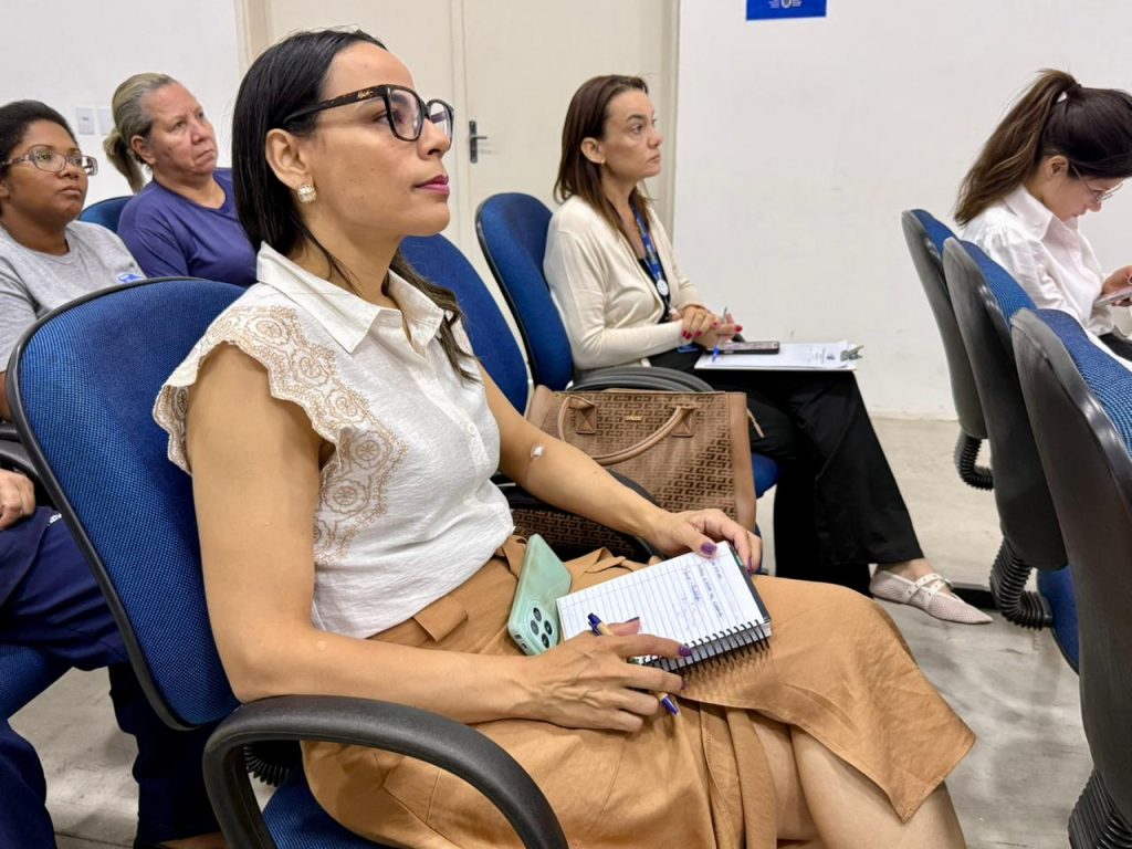 Fotografia em close lateral de uma mulher sentada em um auditório. Ela tem pele clara, cabelos pretos lisos e usa óculos de armação escura. Veste uma blusa branca sem mangas com detalhes bordados e uma saia bege. Ela segura uma caneta e um bloco de notas aberto no colo, olhando atentamente para a frente, em direção ao palestrante (que não aparece na imagem). Ao fundo, outras mulheres aparecem sentadas em poltronas azuis, também prestando atenção.