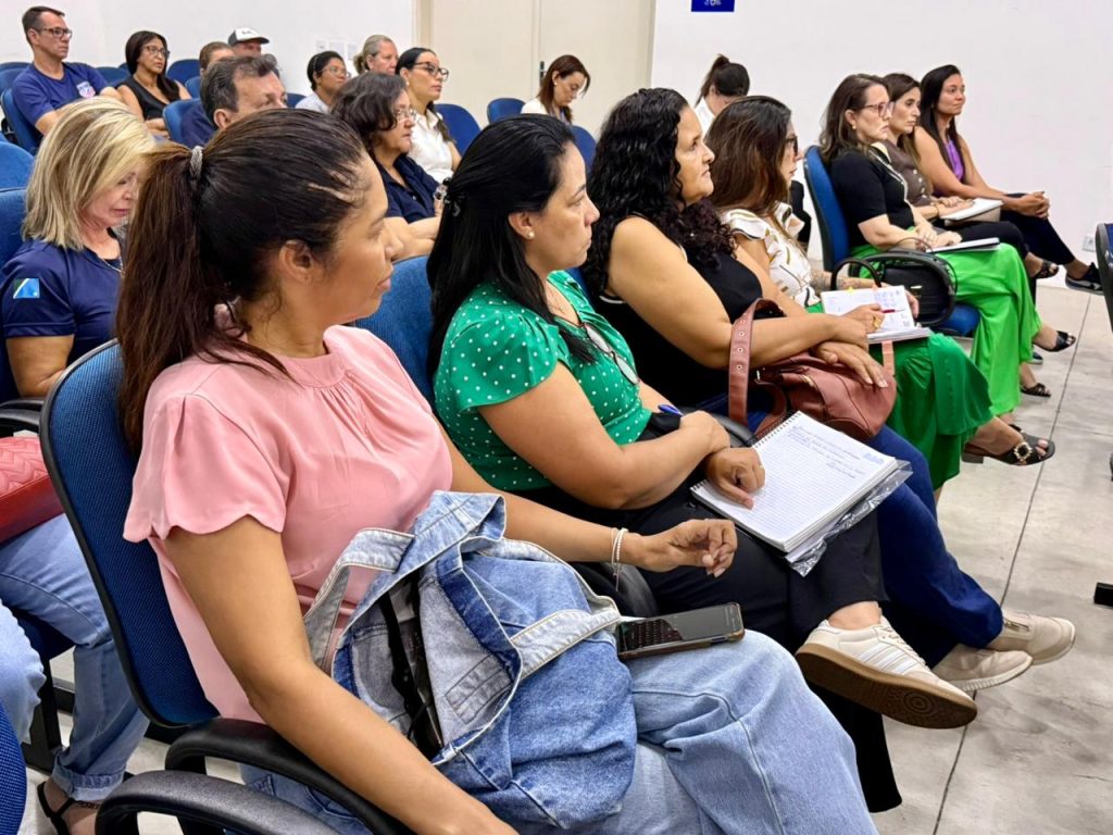 Fotografia lateral mostrando uma fileira de participantes sentados em poltronas azuis. Em primeiro plano, uma mulher de perfil, com cabelos castanhos presos em um rabo de cavalo, usa uma blusa rosa claro e calça jeans. Ao lado dela, outras mulheres aparecem em sequência; uma delas veste blusa verde com bolinhas brancas e segura um caderno. A plateia é composta majoritariamente por mulheres, todas voltadas para o lado direito da foto, indicando que assistem à apresentação.