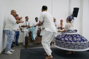Um grupo de pessoas em uma celebração cultural de matriz africana. Ao centro, um homem e uma mulher dançam. A mulher veste uma saia longa e volumosa, estampada em tons de azul, branco e vermelho, que gira com o movimento; ela usa uma blusa de renda branca e um turbante colorido. Os músicos estão ao fundo e à esquerda, tocando tambores (atabaques) de madeira cobertos com tecidos listrados em azul e branco, e um agogô. A maioria veste roupas brancas. O ambiente tem paredes brancas e um tapete escuro no chão.