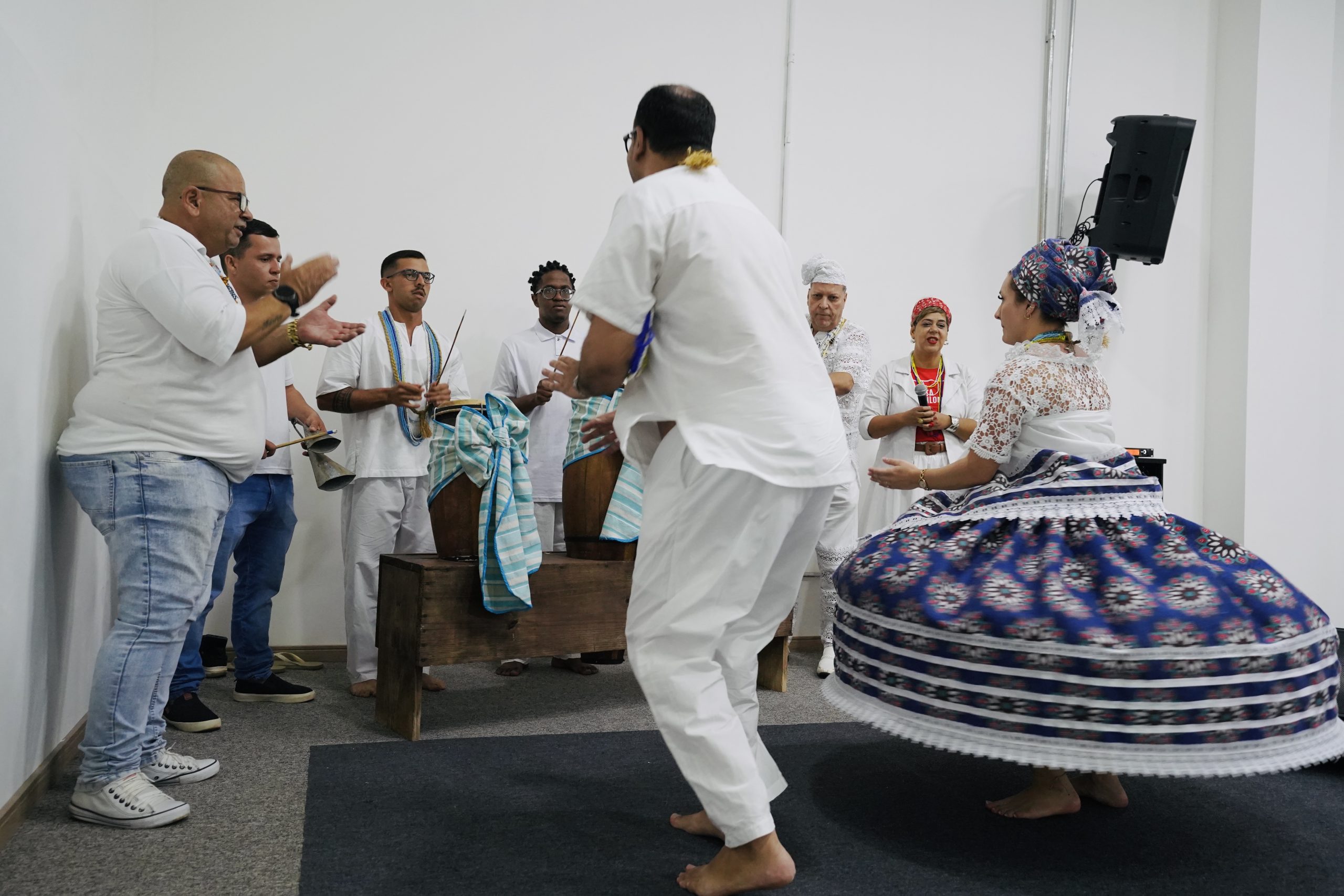 Um grupo de pessoas em uma celebração cultural de matriz africana. Ao centro, um homem e uma mulher dançam. A mulher veste uma saia longa e volumosa, estampada em tons de azul, branco e vermelho, que gira com o movimento; ela usa uma blusa de renda branca e um turbante colorido. Os músicos estão ao fundo e à esquerda, tocando tambores (atabaques) de madeira cobertos com tecidos listrados em azul e branco, e um agogô. A maioria veste roupas brancas. O ambiente tem paredes brancas e um tapete escuro no chão.
