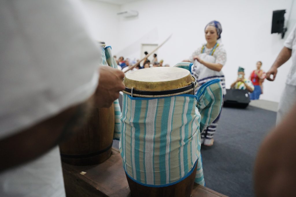 Em primeiro plano, as mãos de um percussionista tocam um atabaque de madeira. O tambor está envolto em um tecido listrado de azul claro e branco, amarrado com um laço lateral. Ao fundo, de forma levemente desfocada, vê-se uma mulher com turbante e vestimentas tradicionais, além de parte da plateia sentada em um auditório. A iluminação é clara e direta.
