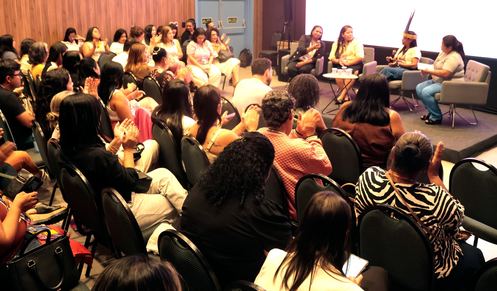 Fotografia de um auditório com plateia predominantemente feminina. No palco, à direita, quatro mulheres participam de uma mesa redonda sentadas em poltronas cinzas. Uma delas usa um cocar indígena. A plateia, em primeiro plano e de costas, está sentada em cadeiras pretas e aplaude o debate. O ambiente possui paredes com revestimento de madeira e iluminação quente.
