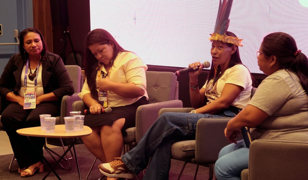 Quatro mulheres sentadas em um painel de discussão. A segunda mulher da direita para a esquerda fala ao microfone; ela é indígena, usa um cocar de penas coloridas e colares tradicionais. As outras integrantes da mesa escutam atentamente. À frente delas, há uma pequena mesa de centro com copos de água. O fundo é composto por um grande telão branco iluminado.
