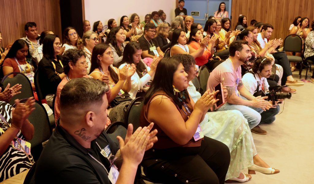 Fotografia tirada da lateral da plateia, mostrando diversas pessoas sentadas e aplaudindo. O público é diverso, composto por homens e mulheres de diferentes etnias. Em destaque no centro, um homem de camiseta rosa e uma mulher com adereços indígenas aplaudem sorridentes. A profundidade de campo mostra o restante do auditório cheio ao fundo.