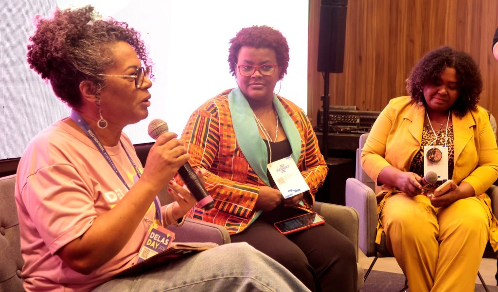 Plano médio de três mulheres negras sentadas em poltronas durante uma palestra. A mulher à esquerda fala ao microfone; ela tem cabelos cacheados presos, usa óculos de grau e camiseta rosa. A mulher ao centro usa óculos vermelhos e um blazer com estampa étnica colorida. A mulher à direita veste um conjunto amarelo vibrante e olha para baixo. Todas usam crachás do Sebrae e do evento "Delas Day".