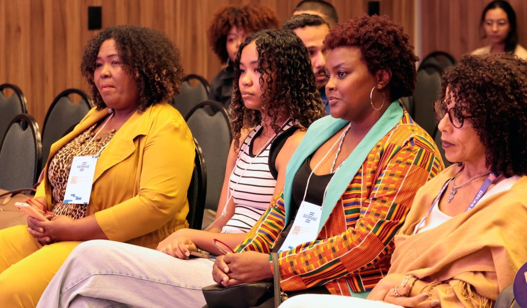 Imagem focada em quatro mulheres sentadas na primeira fila da plateia, vistas de perfil. A segunda mulher da direita para a esquerda ganha destaque: ela possui cabelos curtos e cacheados, usa óculos e um blazer com padrões geométricos em tons de laranja, amarelo e preto. Elas demonstram uma expressão de atenção e seriedade enquanto acompanham o evento.
