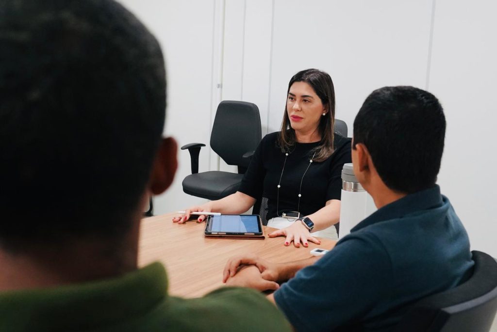Uma mulher de blusa preta está sentada à cabeceira de uma mesa de reunião, gesticulando enquanto fala com dois homens, que aparecem de costas em primeiro plano. Ela utiliza um tablet sobre a mesa. O ambiente tem paredes brancas e cadeiras de escritório pretas.