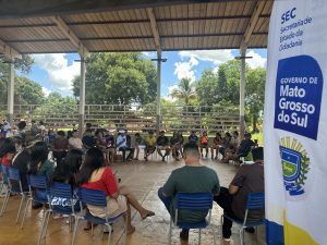 Fotografia em plano aberto de uma reunião sob a estrutura de uma quadra coberta. Dezenas de pessoas, em sua maioria jovens indígenas, estão sentadas em cadeiras azuis dispostas em um grande círculo. Ao fundo, árvores e um céu azul com nuvens brancas. À direita, em primeiro plano, destaca-se um banner branco com o brasão do estado e os dizeres: "SEC - Secretaria de Estado da Cidadania" e "GOVERNO DE Mato Grosso do Sul".