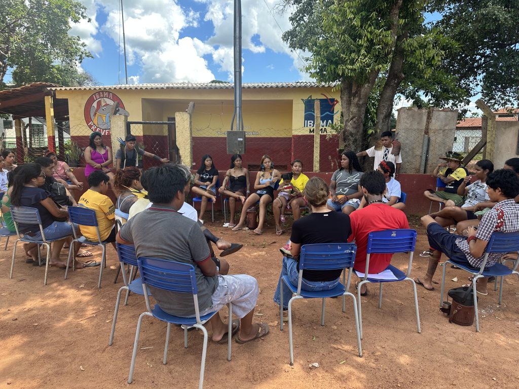 Fotografia em plano aberto de uma reunião ao ar livre. Cerca de vinte pessoas, predominantemente jovens indígenas, estão sentadas em um grande círculo de cadeiras azuis sobre um chão de terra batida. No fundo, há uma construção amarela e vermelha com logotipos do IBAMA e PREVFOGO. Algumas pessoas usam adereços tradicionais, como cocares de penas. O ambiente é arborizado e o céu está parcialmente nublado.