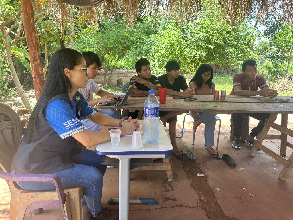 Fotografia de uma reunião de trabalho sob uma cobertura de palha (quiabo). Em primeiro plano, à esquerda, uma mulher jovem de óculos e cabelos longos veste um colete azul com o logotipo "SESAI" (Secretaria de Saúde Indígena). Ela está sentada à mesa com outros cinco jovens indígenas; um deles utiliza um notebook. Sobre a mesa rústica de madeira, há garrafas de água, copos e latas de refrigerante. Ao fundo, vê-se uma área verde com árvores.