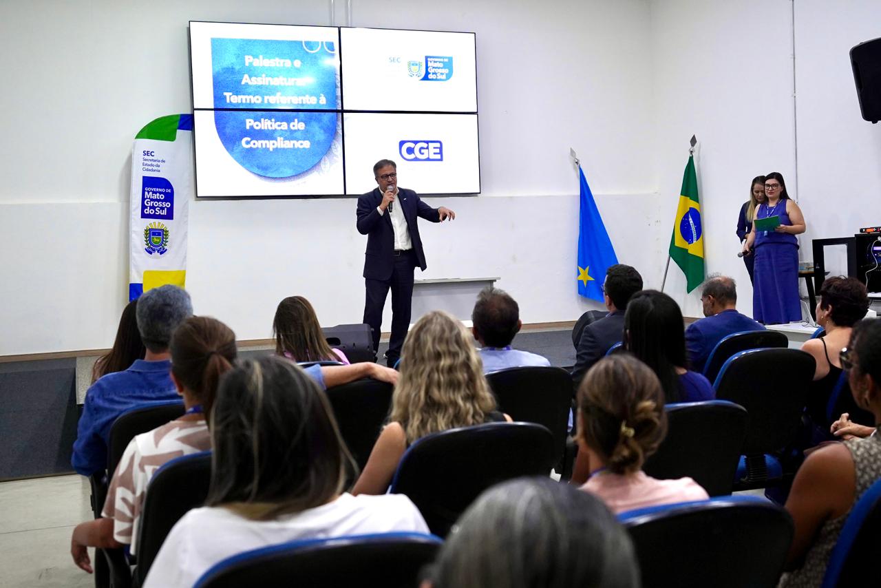 Foto em plano aberto mostrando o auditório durante o evento. O palestrante está ao centro, no palco, gesticulando enquanto fala ao microfone. Em primeiro plano, vemos as costas de várias pessoas sentadas em poltronas azuis, assistindo à apresentação. Ao fundo, o painel digital exibe informações sobre a Política de Compliance.