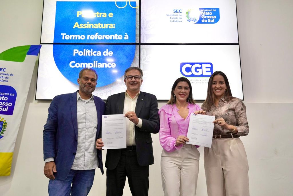 Fotografia de quatro pessoas em pé, posando para a foto em frente ao painel digital do evento. Dois homens e duas mulheres seguram, em dupla, folhas de papel (o termo assinado). O homem ao centro-esquerda e a mulher ao centro-direita (vestindo uma blusa rosa) sorriem para a câmera. Todos estão vestidos de forma social/casual.