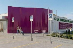 Imagem de uma renderização 3D da entrada principal da "Casa da Mulher Brasileira". Em destaque, uma grande parede curva na cor roxa vibrante. À direita, a fachada branca exibe o nome do local em letras verdes aplicadas. Há rampas de acesso com corrimãos metálicos e placas de sinalização com o símbolo internacional de acessibilidade (cadeirante). Na calçada, uma mulher em uma cadeira de rodas é conduzida por outra mulher. Ao fundo, um grupo de mulheres conversa próximo a uma área de jardim. O ambiente é ensolarado e amplo.