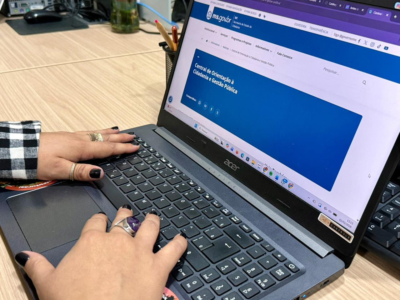 Foto das mãos de uma mulher com anéis e unhas pintadas de preto, digitando em um notebook. Na tela, está aberto o site oficial do Governo de Mato Grosso do Sul, na página da "Central de Orientação à Cidadania e Gestão Pública". Ao fundo, mesa de trabalho com itens de escritório.
