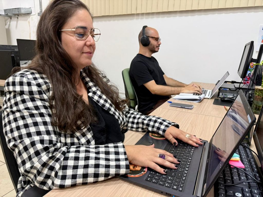 Uma mulher de óculos e blazer xadrez preto e branco trabalha em um notebook em uma mesa de escritório. Ao lado dela, um homem de camiseta preta e fones de ouvido também utiliza um computador. O ambiente é um escritório iluminado e organizado.