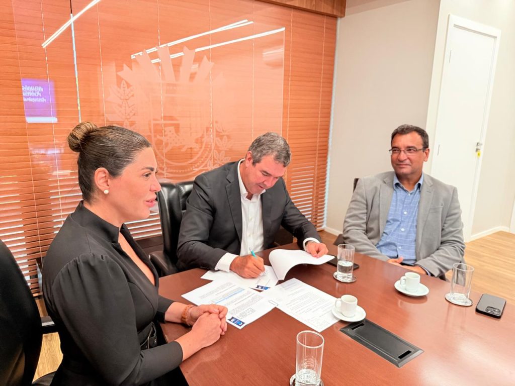 Close em três pessoas à cabeceira da mesa. Um homem de terno escuro está inclinado, assinando um documento com uma caneta azul. À esquerda, uma mulher de preto observa e, à direita, um homem de paletó cinza sorri.