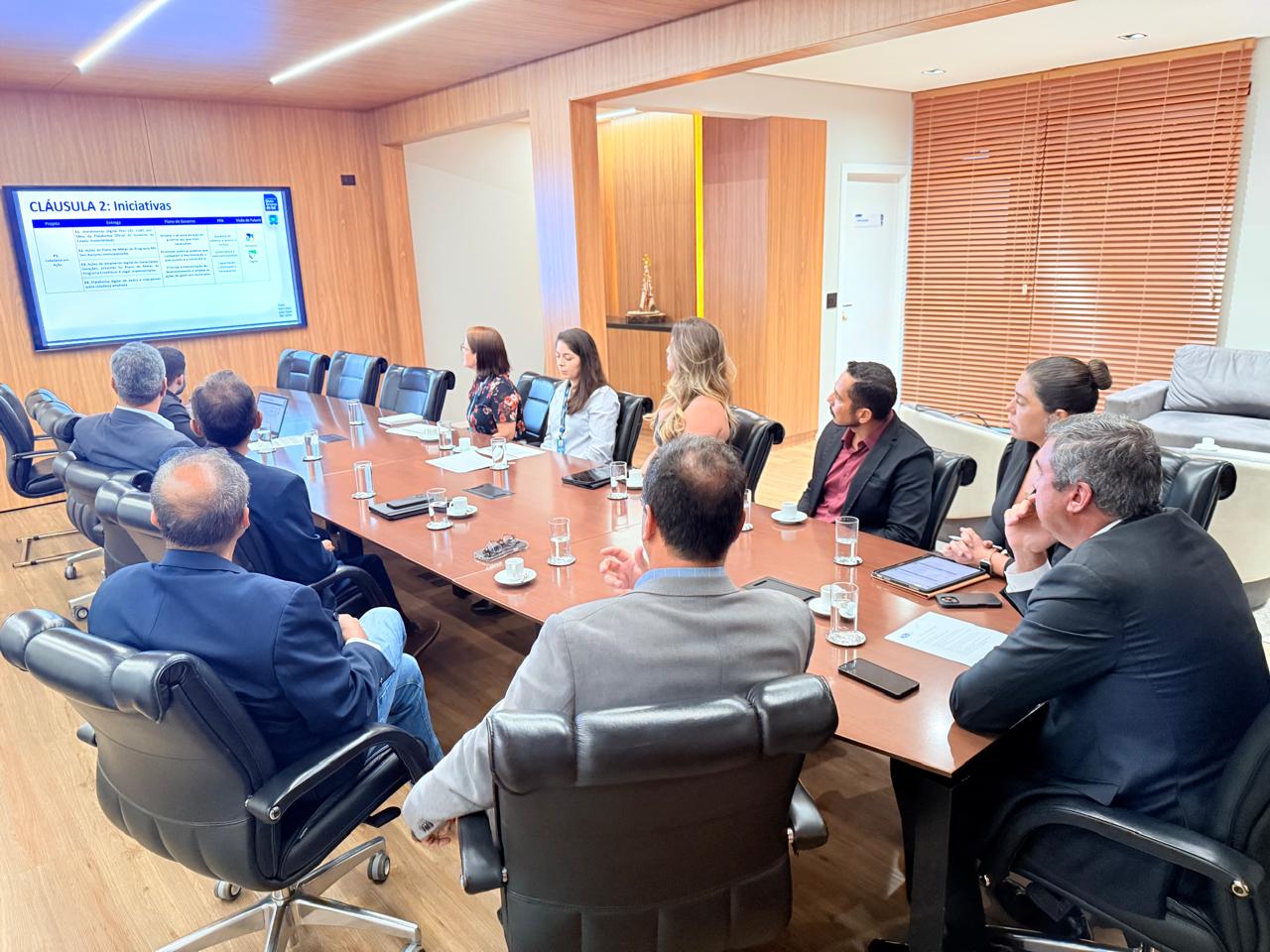 Vista lateral de uma sala de reuniões com várias pessoas sentadas ao redor de uma mesa de madeira. Ao fundo, uma tela de TV exibe um slide azul e branco intitulado "CLÁUSULA 2: Iniciativas", contendo uma tabela com metas de governo.